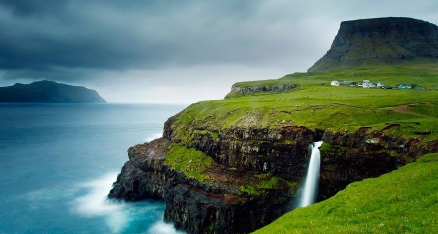 Das Dorf Gásadalur unter dem Heinanova-Berg, mit Wasserfall über das Kliff in den Atlantik, Färöer-Inseln